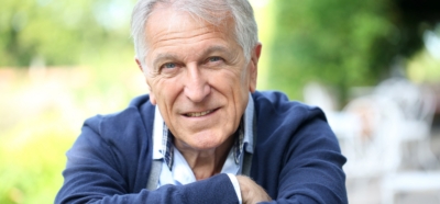 Homme senior souriant, assis en extérieur, regard chaleureux et bienveillant, symbole de confiance et d’engagement.