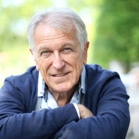 Homme senior souriant, assis en extérieur, regard chaleureux et bienveillant, symbole de confiance et d’engagement.
